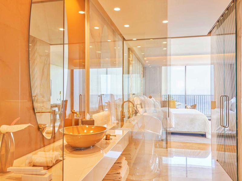 Moderní koupelna se skleněnou zdí, dvojnásobnými umyvadly a výhledem na ložnici s výhledem na moře.