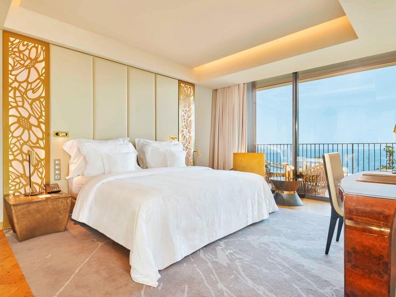 Moderní hotelový pokoj s velkou postelí, kobercem a okny od podlahy ke stropu s výhledem na moře.