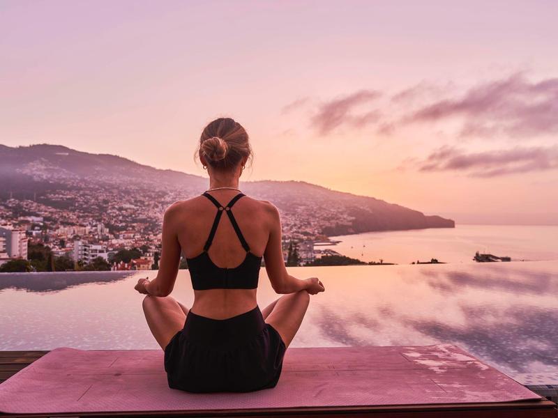 Žena meditujíc na jógové podložce s výhledem na klidnou vodu a město při západu slunce.
