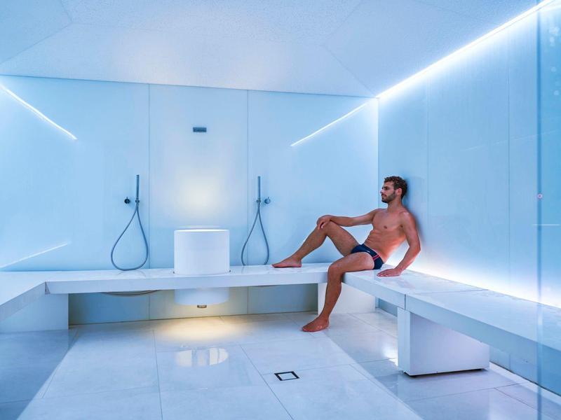 Muž sedící v moderní bílé wellness místnosti s lavicemi a sprchami, obklopený ambientním osvětlením.