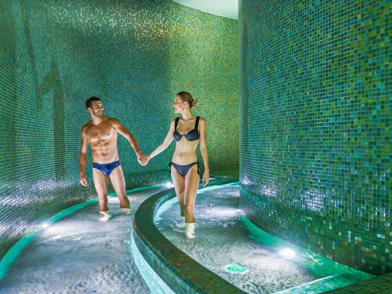 Pár jdoucí za ruku osvětlenou spa oblastí s mozaikovými dlaždicemi a vodou.