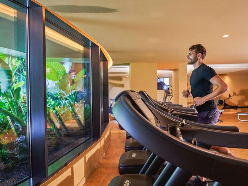 Muž běží na běžeckém pásu v moderní posilovně s velkým panoramatickým oknem a výhledem na rostliny.
