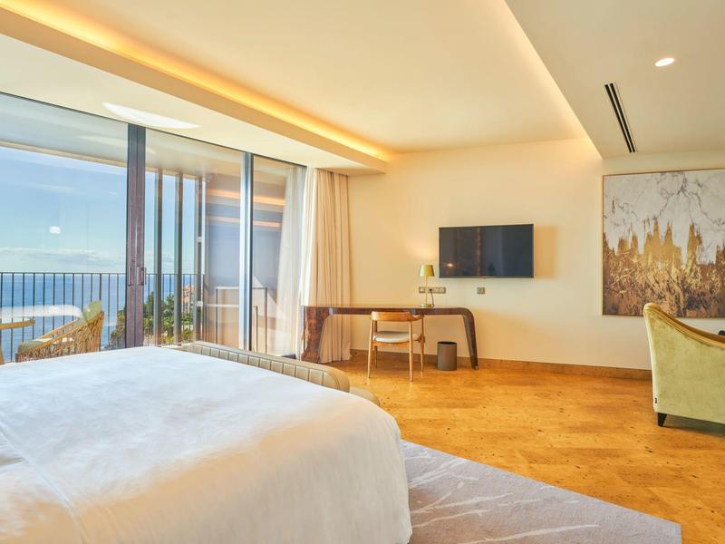 Moderní hotelový pokoj s výhledem na moře, velkou postelí a posezením.