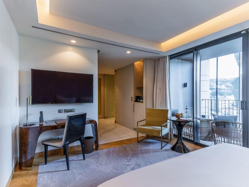 Moderní hotelový pokoj s TV, židlemi, stolem a balkonem s výhledem na město.