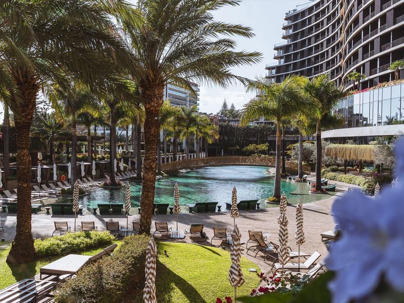 Luxusní hotelový bazén obklopený palmami a lehátky vedle moderní vysoké budovy.