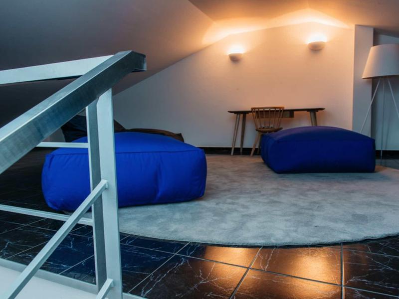 Cozy attic space with blue beanbags, a round carpet, wooden table, and lamp under soft lighting.
