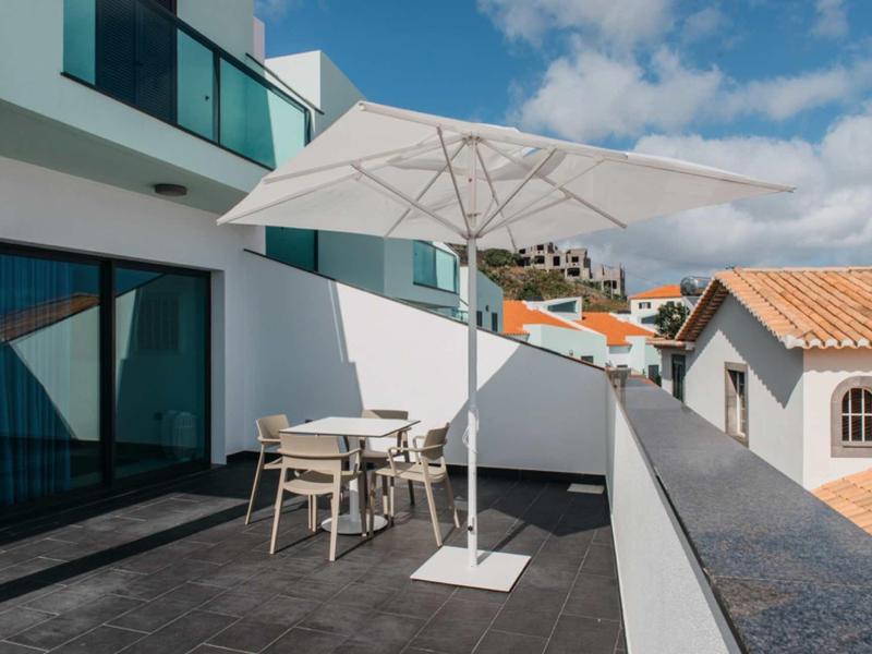 Spacious balcony with table, chairs, and umbrella overlooking rooftops under blue sky.