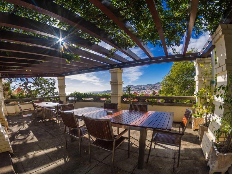 Overdekt terras met tafels en stoelen, uitzicht op tuin en heuvels in zonlicht.