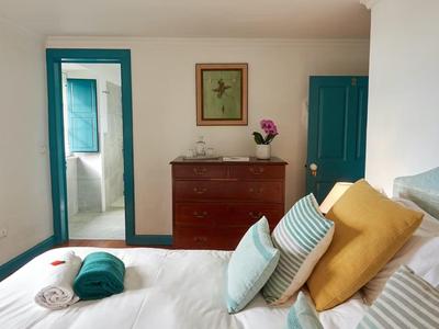 Světlo zařízená ložnice s bílou postelí, barevnými polštáři a hnědou komodou u modrých dveří.