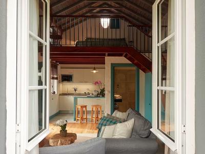 Výhled přes otevřené okno do útulného obývacího pokoje s pohovkou, dřevěným stolem a otevřenou kuchyní.