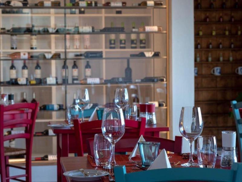 Restaurace s červenými a modrými židlemi a skleněnou vitrínou plnou lahví vína.