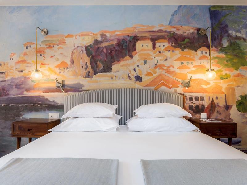 Modern hotelkamer met tweepersoonsbed en muurschildering van een Middellandse Zeestads.