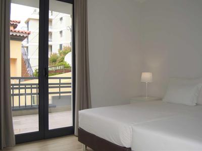 Licht, eenvoudig ingericht hotelkamer met twee eenpersoonsbedden en balkon met uitzicht op andere gebouwen.