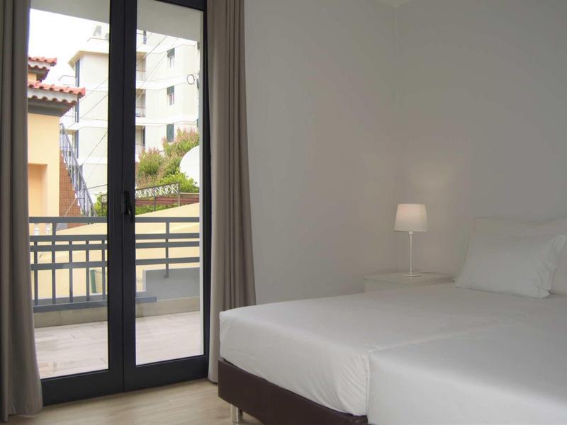 Licht, eenvoudig ingericht hotelkamer met twee eenpersoonsbedden en balkon met uitzicht op andere gebouwen.