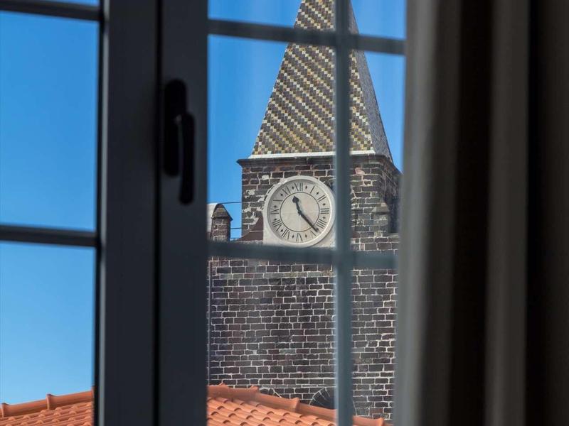 Uitzicht vanuit een hotelraam op een klokkentoren achter een rood dak bij heldere lucht.