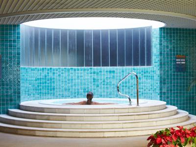 Persona si rilassa in una vasca idromassaggio rotonda sotto una grande apertura sul soffitto, circondata da piastrelle turchesi.