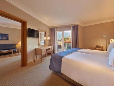 Camera d'hotel luminosa con letto grande, balcone e area soggiorno adiacente.