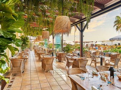 Ristorante all'aperto con tavoli apparecchiati e piante sotto una pergola al sole.