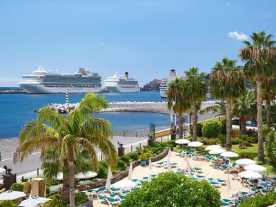 Blick auf eine Küste mit Palmen, Liegestühlen und zwei Kreuzfahrtschiffen im Hintergrund.