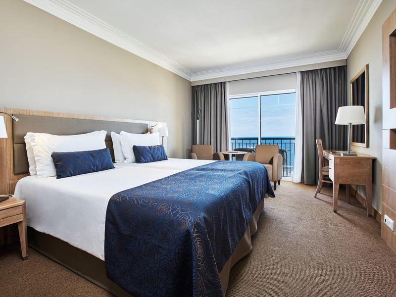 Camera d'hotel luminosa con letto matrimoniale, copriletto blu e balcone con vista mare.