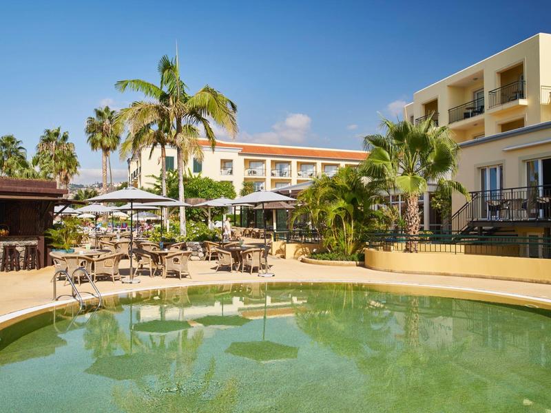 Struttura alberghiera con piscina, palme e ombrelloni in una giornata soleggiata.