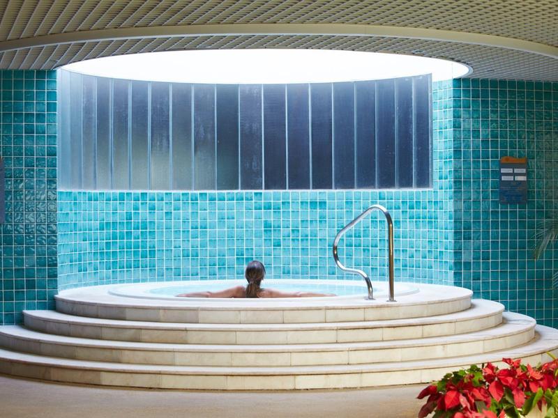 Persona si rilassa in una vasca idromassaggio rotonda sotto una grande apertura sul soffitto, circondata da piastrelle turchesi.
