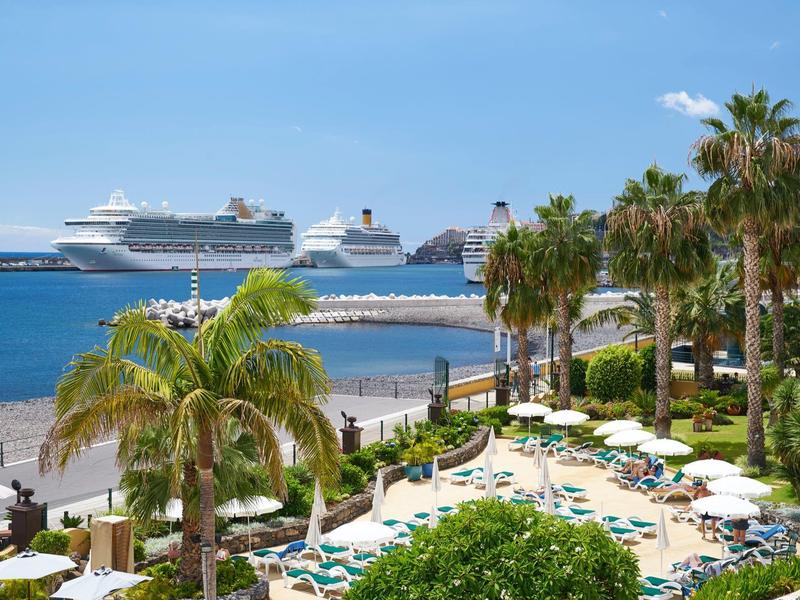 Resort sulla spiaggia con palme, lettini e vista su due navi da crociera in mare.
