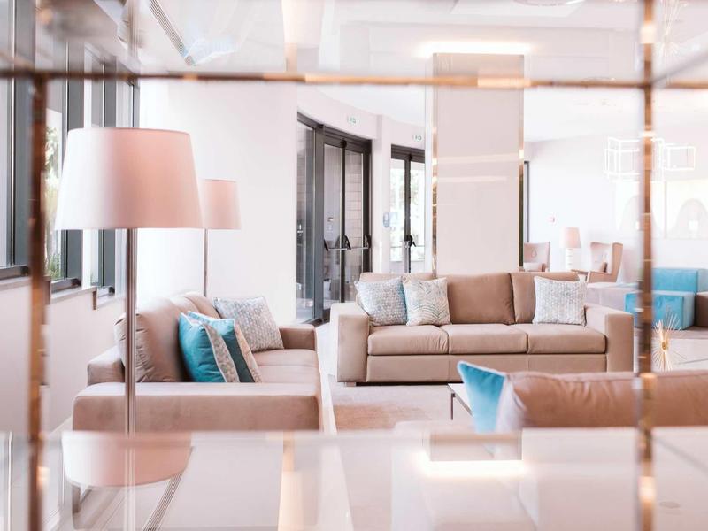 Helles Wohnzimmer mit beigen Sofas, blauen Kissen und mehreren Lampen vor großen Fenstern.