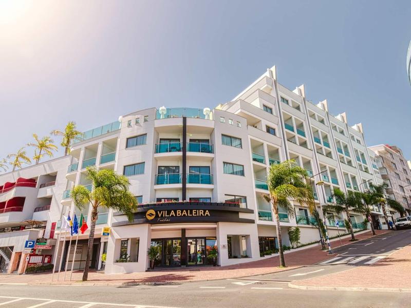 Modernes, weißes Eckgebäude mit großen Glasfenstern und Palmen entlang der Straße bei klarem Himmel.