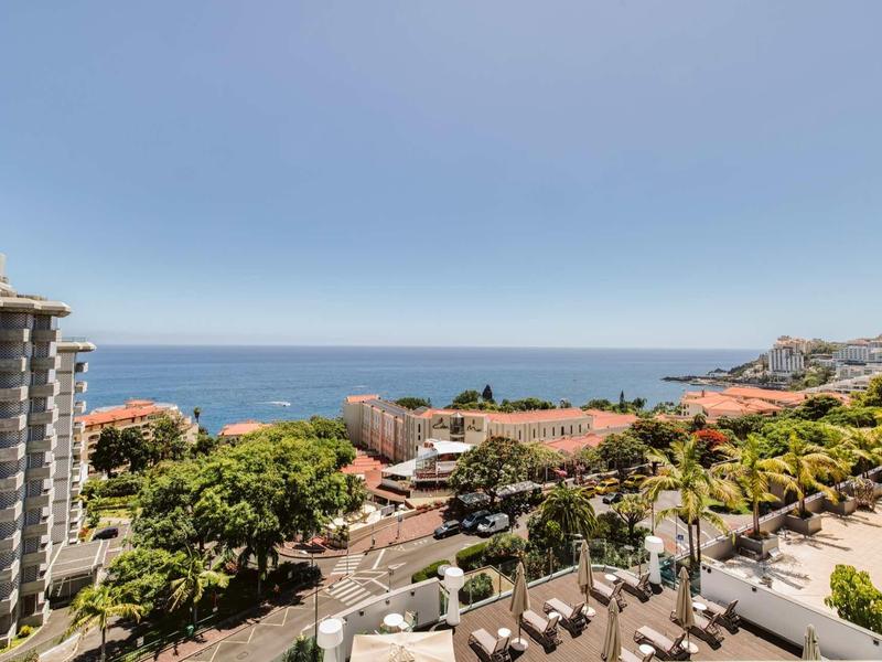 Blick auf Stadt mit grünen Bäumen, Wohnhäusern und Meer am Horizont unter blauem Himmel.