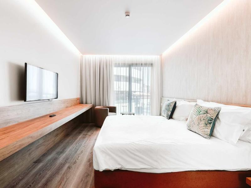 Helles Schlafzimmer mit großem Bett, weißen Vorhängen, Holzboden und Wandregal mit Fernseher.