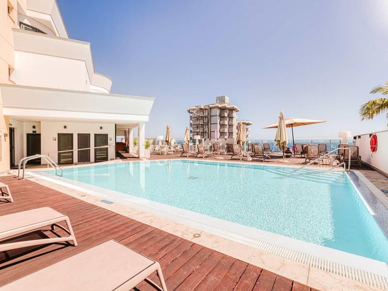 Ein großer Pool mit hellblauem Wasser auf Holzdeck, umgeben von Liegen und Sonnenschirmen bei Sonnenschein.