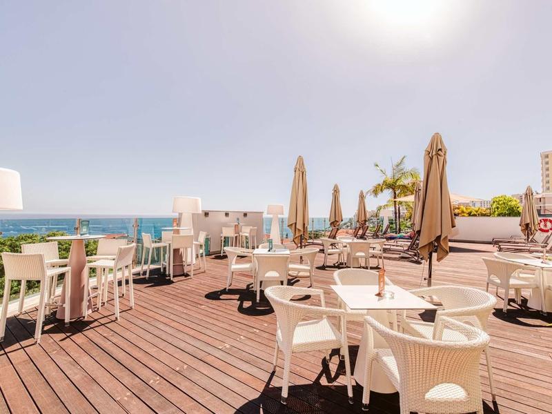Dachterrasse mit weißen Sitzmöbeln, Sonnenschirmen, Holzboden und Meerblick bei sonnigem Himmel.