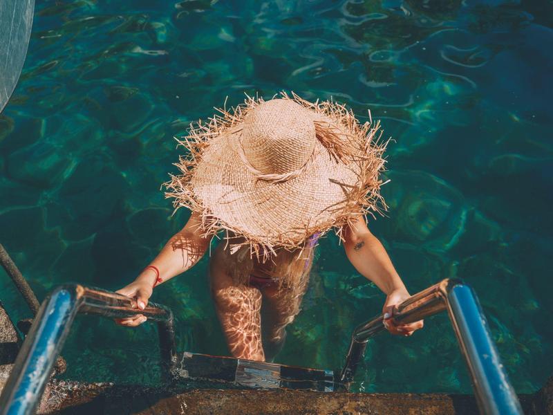 Persona con cappello di paglia sale una scala dall'acqua limpida blu-verde di una piscina o mare.