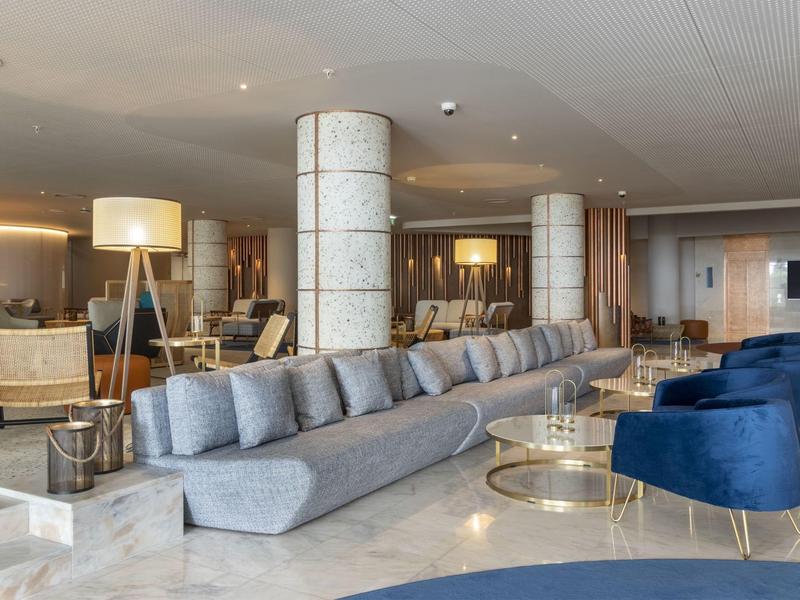 Lobby moderna di un hotel con divano grigio, poltrone blu e tavolini dorati.