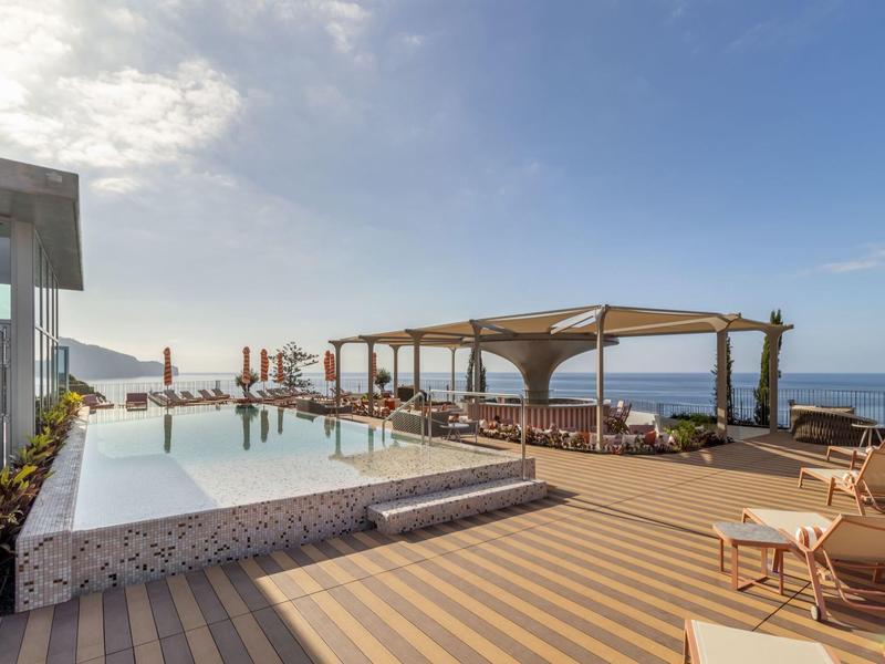 Area piscina moderna con lettini, pergolato e vista mare sotto un cielo azzurro.