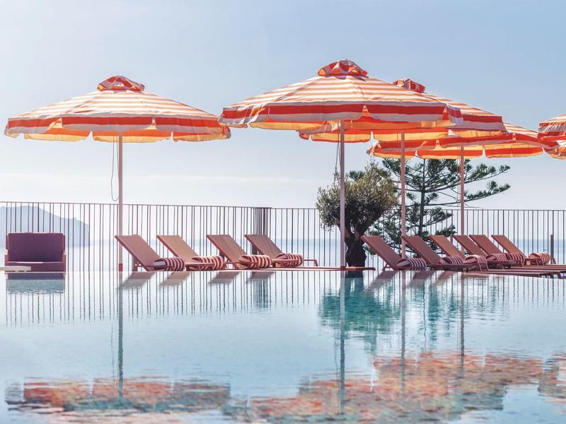 Area piscina con ombrelloni rossi e lettini accanto a una piscina a sfioro limpida.