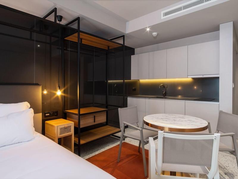 Camera d'hotel moderna con letto, area salotto e angolo cottura aperto.