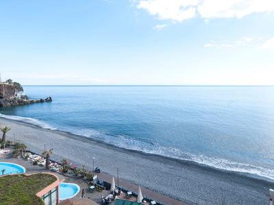 Klares blaues Meer trifft auf grauen Sandstrand mit Palmen und blauem Pool im Vordergrund.