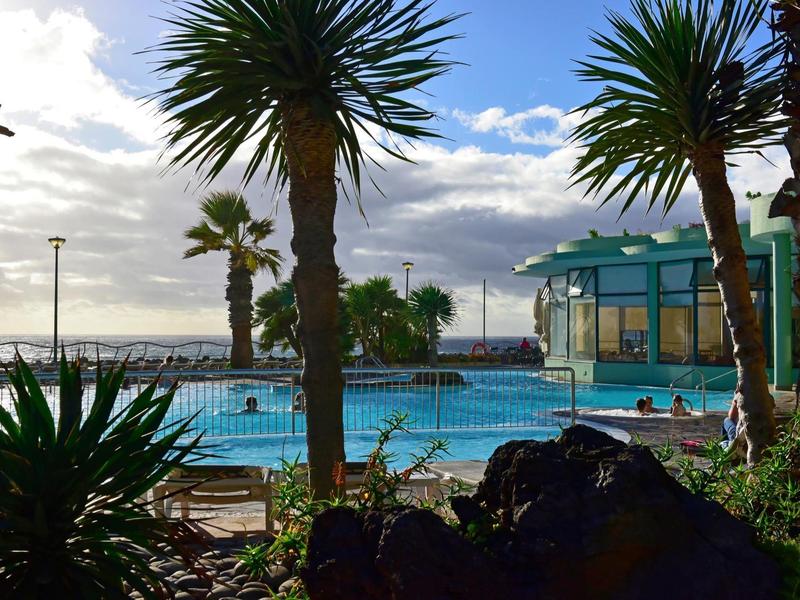Vista di una piscina con palme e un edificio contro un orizzonte marino nuvoloso.