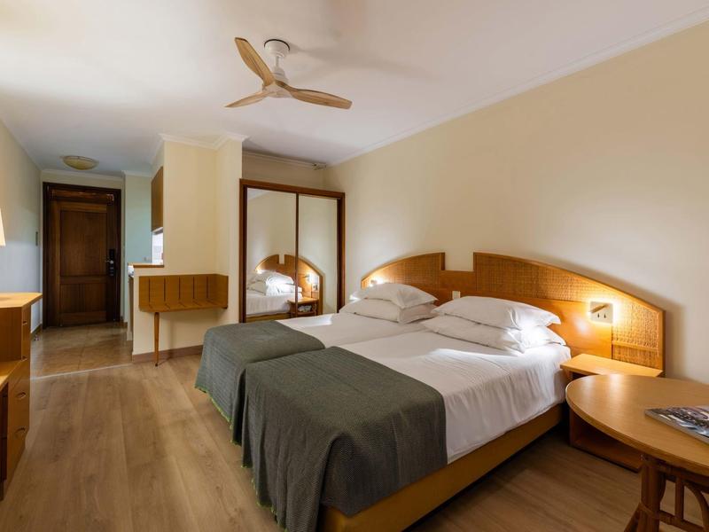 Camera d'hotel luminosa con letto matrimoniale, mobili in legno e ventilatore a soffitto.