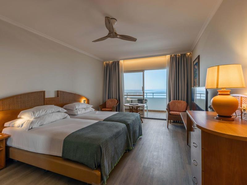 Camera d'hotel luminosa arredata con letto matrimoniale, poltrone e balcone con vista mare.