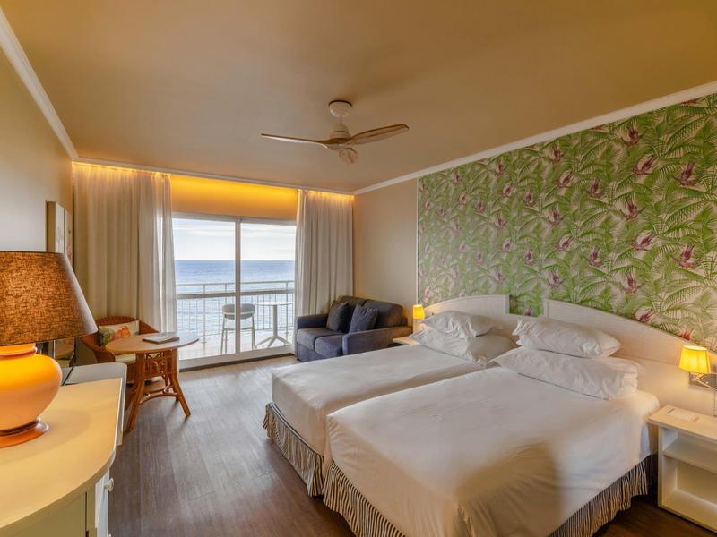 Camera d'hotel con due letti singoli, parete a motivo verde e balcone con vista mare.