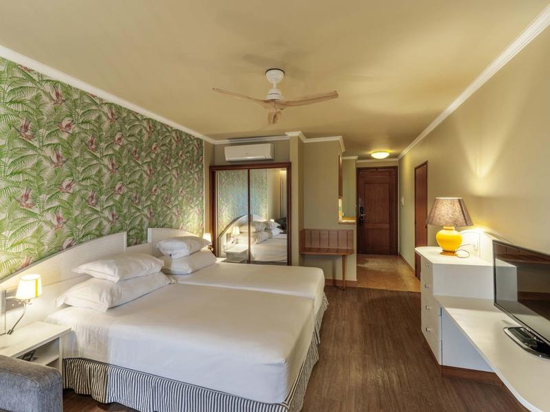 Camera d'hotel luminosa con due letti, carta da parati floreale, ventilatore a soffitto e TV.