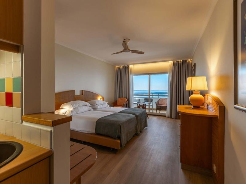 Camera d'hotel luminosa con letto matrimoniale, zona salotto e vista mare attraverso grandi finestre.