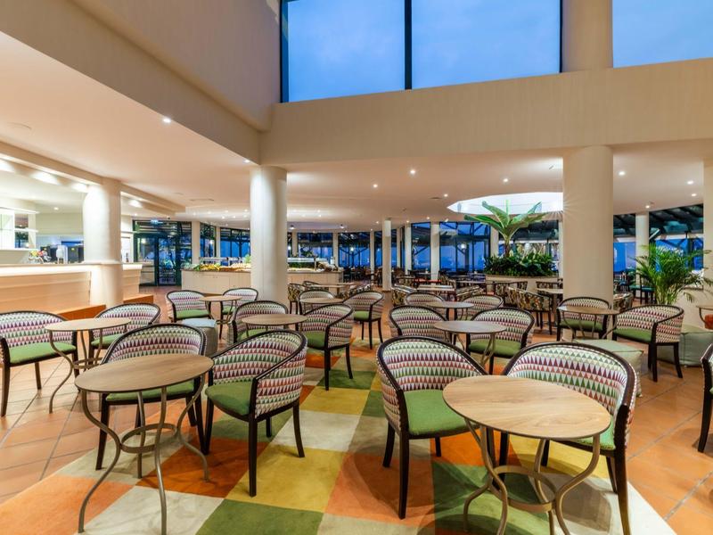 Ampia sala da pranzo luminosa con sedie in rattan, tavoli, grandi finestre e pavimento piastrellato.