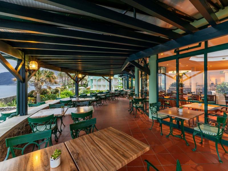 Terrazza coperta con tavoli e sedie con vista sul mare e le montagne al tramonto.