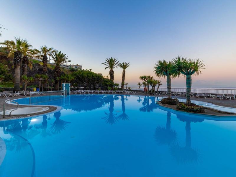 Grande piscina all'aperto circondata da palme, vista sul mare al tramonto.