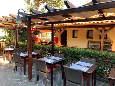 Außenterrasse eines Restaurants mit Holztischen, Stühlen und Pergola an sonnigem Tag.