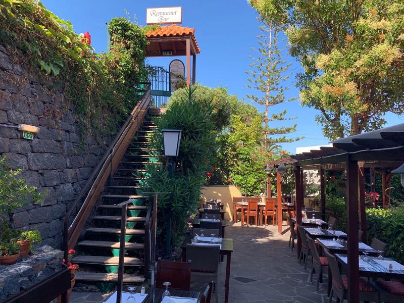 Area esterna soleggiata con scale, pareti coperte di piante e tavoli apparecchiati in un ristorante.